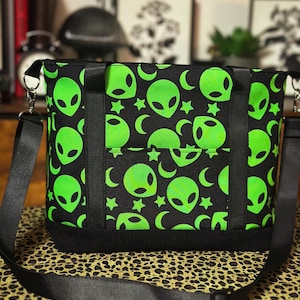 May include: Black tote bag with a neon green alien face pattern, stars, and crescent moons. The bag has black straps and a black bottom. The bag has a black adjustable shoulder strap.