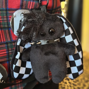May include: A black plush rabbit keychain with a spiked collar, long ears featuring a black and white checkered pattern, and a tuft of black hair. The plush toy is attached to a red, black, and blue plaid bag with a circular pin showing a person.