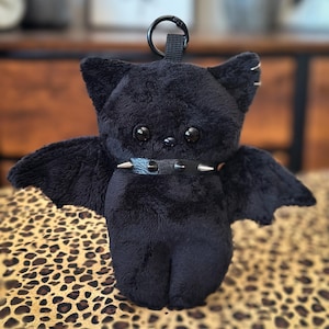 Könnte beinhalten: Ein schwarzes Plüschtier in Form einer Fledermaus mit großen, runden Augen und kleinen, spitzen Ohren. Die Fledermaus hat ausgebreitete Flügel und einen schwarzen Kragen mit silbernen Stacheln. Oben ist eine schwarze Schlaufe zum Aufhängen angebracht.