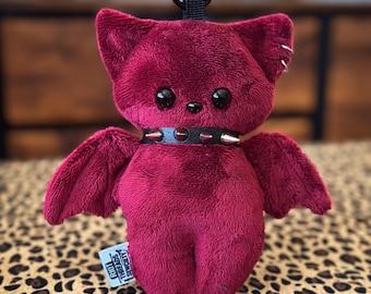 May include: Plush burgundy cat-like creature with black eyes, small wings, and a studded collar. The toy has a keychain loop and a small tag that reads "Idiot Threads Society". The background is blurred, with a leopard print surface.