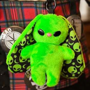 May include: A neon green plush alien bunny keychain. The bunny has large black eyes and a pink nose. The inside of the ears are black with white stars and green alien heads. The keychain is attached to a black ring.
