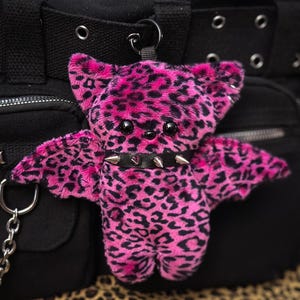 Può includere: Un portachiavi a forma di pipistrello in peluche con motivo leopardato rosa acceso e nero. Il pipistrello presenta due occhi in plastica nera, un piccolo naso nero e un collare in similpelle nera decorato con borchie metalliche argentate.
