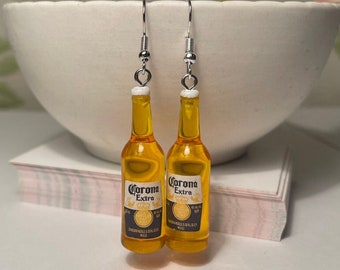 May include: A pair of silver dangle earrings featuring miniature Corona Extra beer bottles. The bottles are yellow and have the Corona Extra logo printed on them.