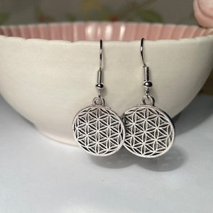 May include: A pair of silver earrings featuring a flower of life design. The earrings are hanging from a white bowl.