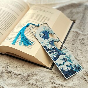 K&ouml;nnte beinhalten: Ein rechteckiger Lesezeichen mit einem blau-wei&szlig;en Wellenmuster und einer blauen Quaste. Das Lesezeichen befindet sich in einem offenen Buch auf einer weichen, strukturierten Oberfl&auml;che.