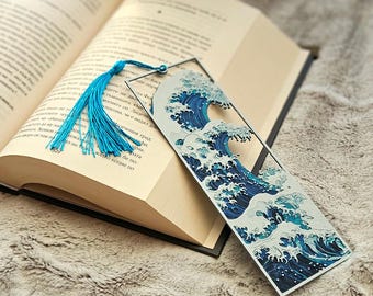 May include: A rectangular bookmark featuring a stylized blue and white wave design, with a matching blue tassel. The bookmark is placed in an open book on a soft, textured surface.