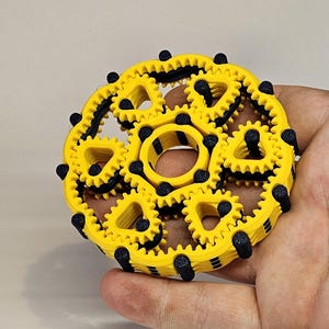 May include: A yellow and black gear toy with multiple interlocking gears. The gears are arranged in a circular pattern and have a black centre.