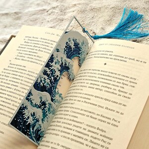 K&ouml;nnte beinhalten: Dekorativer Lesezeichen mit blau-wei&szlig;em Wellenmuster und passendem blauen Quastenanh&auml;nger. Das Lesezeichen steckt in einem Buch mit Text auf den Seiten.