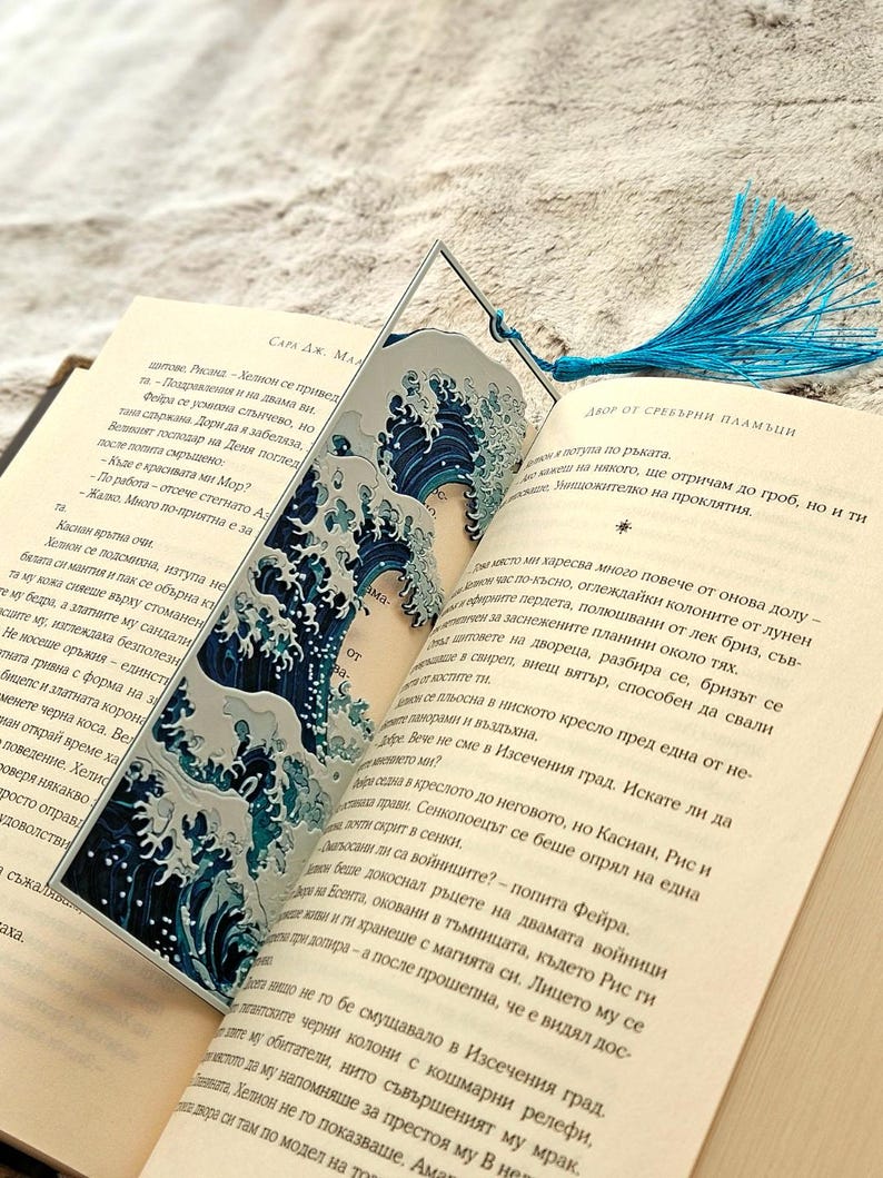 K&ouml;nnte beinhalten: Dekorativer Lesezeichen mit blau-wei&szlig;em Wellenmuster und passendem blauen Quastenanh&auml;nger. Das Lesezeichen steckt in einem Buch mit Text auf den Seiten.
