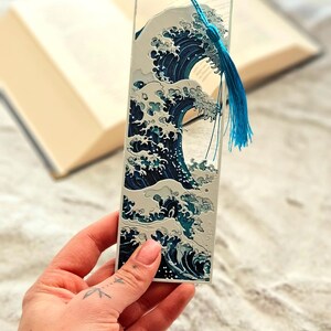 K&ouml;nnte beinhalten: Rechteckiger Lesezeichen mit einem stilisierten Wellenmuster in Blau- und Wei&szlig;t&ouml;nen. Das Lesezeichen hat eine blaue Quaste. Das Design ist von japanischer Kunst inspiriert.