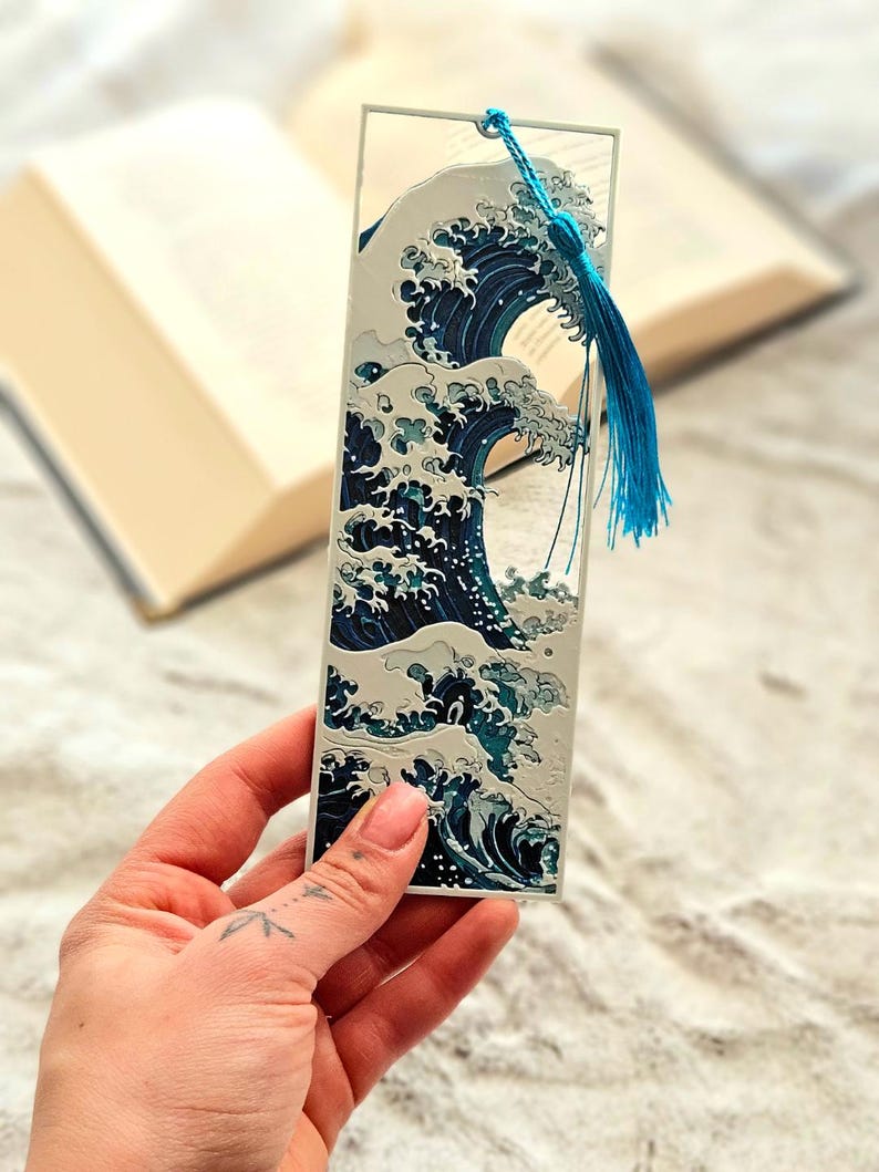 K&ouml;nnte beinhalten: Rechteckiger Lesezeichen mit einem stilisierten Wellenmuster in Blau- und Wei&szlig;t&ouml;nen. Das Lesezeichen hat eine blaue Quaste. Das Design ist von japanischer Kunst inspiriert.