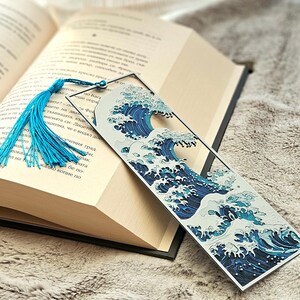 K&ouml;nnte beinhalten: Ein Lesezeichen mit einem blau-wei&szlig;en Wellenmuster und einer blauen Quaste. Das Lesezeichen befindet sich in einem offenen Buch mit cremefarbenen Seiten. Das Design ist von japanischer Kunst inspiriert.