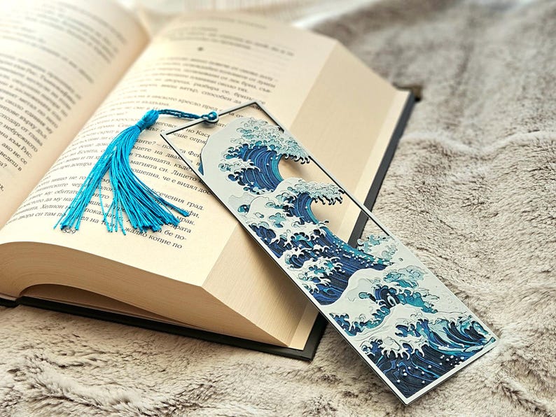 K&ouml;nnte beinhalten: Ein Lesezeichen mit einem blau-wei&szlig;en Wellenmuster und einer blauen Quaste. Das Lesezeichen befindet sich in einem offenen Buch mit cremefarbenen Seiten. Das Design ist von japanischer Kunst inspiriert.