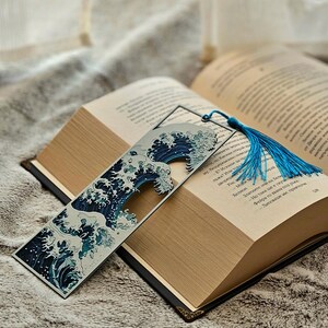 K&ouml;nnte beinhalten: Ein Lesezeichen mit blau-wei&szlig;em Wellenmuster, das in einem offenen Buch liegt. Das Lesezeichen hat eine blaue Quaste. Das Buch hat einen schwarzen Einband und goldfarbene Kanten. Das Buch liegt auf einer weichen, strukturierten Oberfl&auml;che.