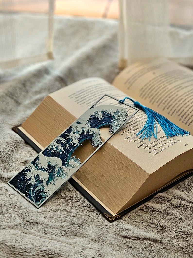 K&ouml;nnte beinhalten: Ein Lesezeichen mit blau-wei&szlig;em Wellenmuster, das in einem offenen Buch liegt. Das Lesezeichen hat eine blaue Quaste. Das Buch hat einen schwarzen Einband und goldfarbene Kanten. Das Buch liegt auf einer weichen, strukturierten Oberfl&auml;che.