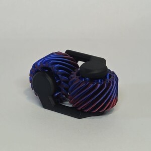 May include: A close-up of a 3D printed gear mechanism with two gears. The gears are blue and red with a black plastic frame.