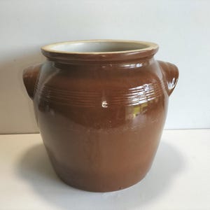 Könnte beinhalten: Ein brauner Keramiktopf mit glänzender Oberfläche. Der Topf hat eine weite Öffnung, zwei seitliche Griffe und horizontale Rillen in der Mitte. Der Innenraum ist hellcremefarben. Dieser Vintage-Küchenartikel eignet sich zur Aufbewahrung oder Präsentation.