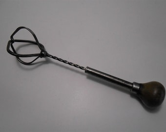 Può includere: Una frusta vintage in metallo con manico in legno. La frusta ha un'asta in metallo attorcigliata e una gabbia di filo metallico nella parte superiore. Il manico è marrone e ha una forma arrotondata.