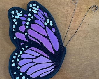 May include: A purple and black butterfly with white dots, made of fabric and wire. The butterfly is attached to a wire that is bent into two loops.