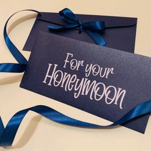 May include: A navy blue card and envelope set, with the words "For your Honeymoon" in white script. A matching blue ribbon is tied in a bow on the envelope and also used as a decorative element.