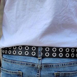 May include: A black leather belt with silver grommets. The belt is worn around the waist of a person wearing blue jeans.