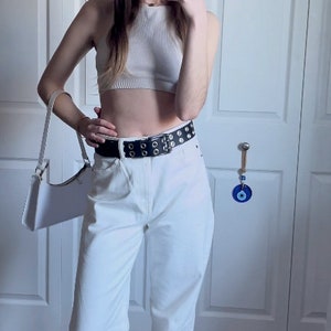 May include: A white crop top with a white belt with silver grommets. The belt is worn over white high-waisted pants.