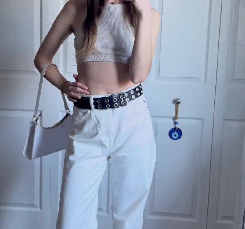 May include: A white crop top with a white belt with silver grommets. The belt is worn over white high-waisted pants.