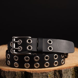 May include: A black leather belt with silver metal eyelets and a double buckle. The belt is rolled up and sitting on a wooden surface.