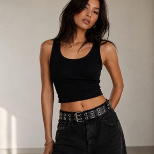 May include: A black tank top with a scoop neckline. The top is paired with black denim jeans and a black belt with silver grommets. The model is wearing a silver bracelet and a necklace.