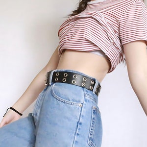 May include: A woman wearing a red and white striped cropped top with a black belt with silver eyelets and blue jeans.