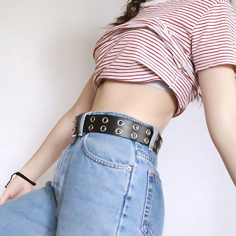 May include: A woman wearing a red and white striped cropped shirt with a black belt with silver grommets and blue jeans.