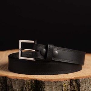 May include: A black leather belt with a silver buckle. The belt is rolled up and sitting on a wooden surface.