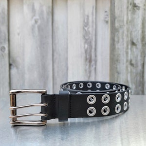 May include: A black leather belt with silver eyelets and a double prong buckle. The belt is on a silver surface.