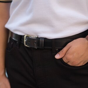 May include: A person wearing a white polo shirt with a black leather belt and a brown leather wristwatch with a white face.