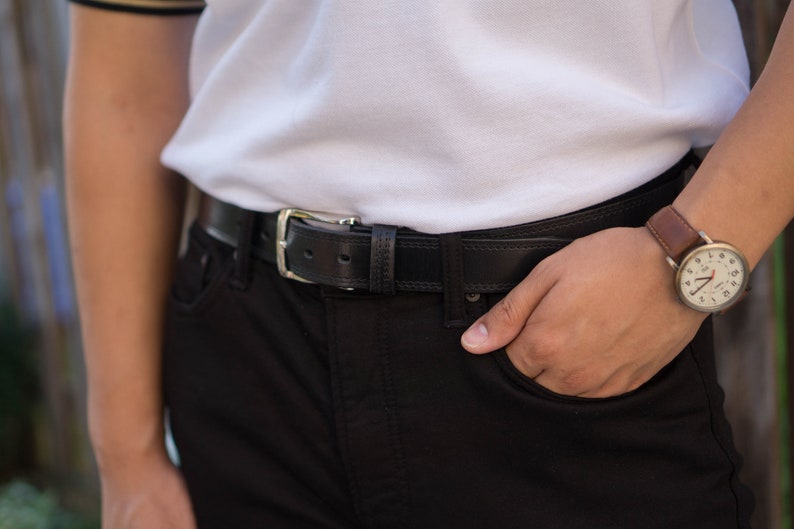 May include: A person wearing a white polo shirt with a black leather belt and a brown leather wristwatch with a white face.
