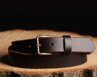 May include: A brown leather belt with a brass buckle. The belt is rolled up and sitting on a wooden surface.