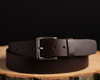 May include: A brown leather belt with a silver buckle. The belt is folded in half and sitting on a wooden surface.