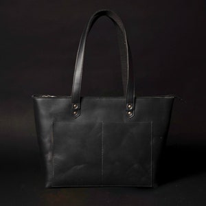 May include: A black leather tote bag with two front pockets and a zip closure. The bag has two top handles.