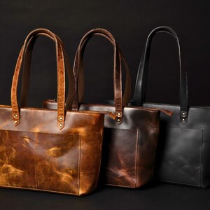 May include: Three leather tote bags in different shades of brown and black. The bags have a zipper closure and a single pocket on the front.