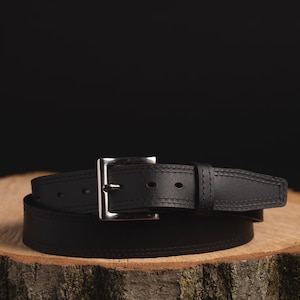 May include: A black leather belt with a silver buckle. The belt is wrapped around a piece of wood.