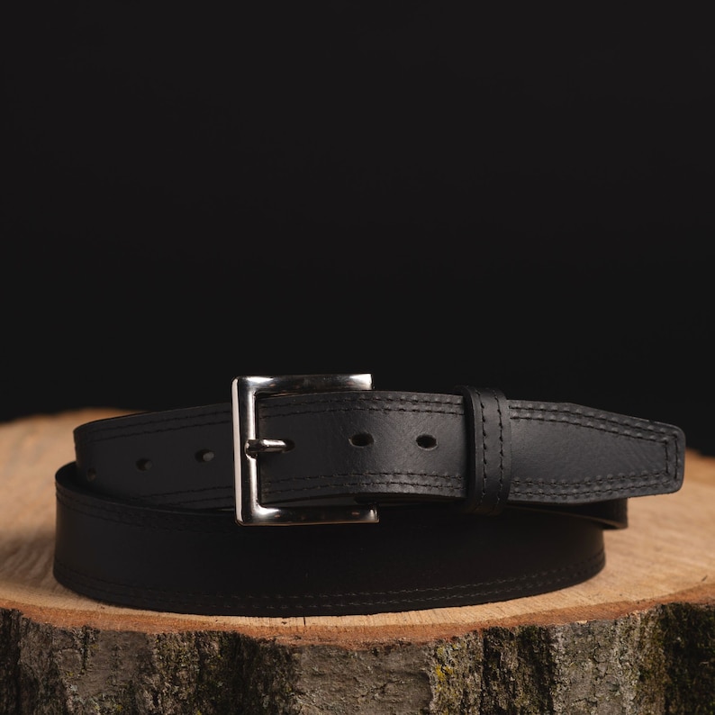 May include: A black leather belt with a silver buckle. The belt is wrapped around a piece of wood.