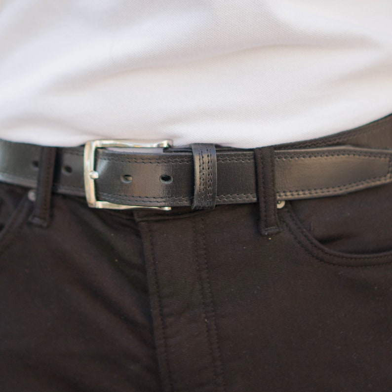 May include: A black leather belt with a silver buckle. The belt is fastened around a pair of black pants.