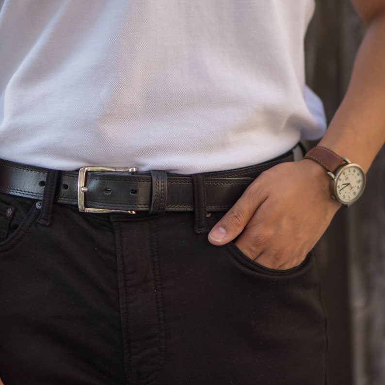 May include: A black leather belt with a silver buckle. The belt is worn around the waist of a person wearing black jeans.