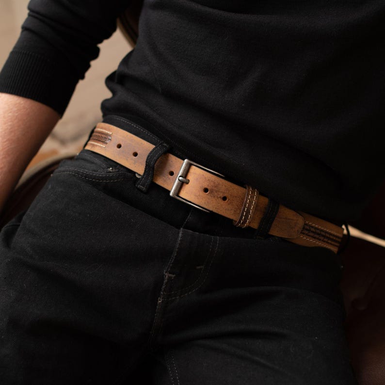 May include: A brown leather belt with a silver buckle is worn over black jeans. The belt has a distressed appearance with visible stitching and dark accents. The belt is approximately 1.5 inches wide.