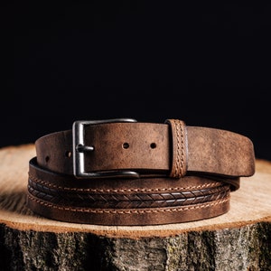 May include: A brown leather belt with a silver-colored buckle. The belt features a woven design detail and is displayed on a wooden surface. The leather has a distressed appearance, and the belt is rolled up.