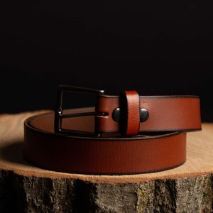 May include: A brown leather belt with a silver buckle. The belt is rolled up and sitting on a wooden surface.