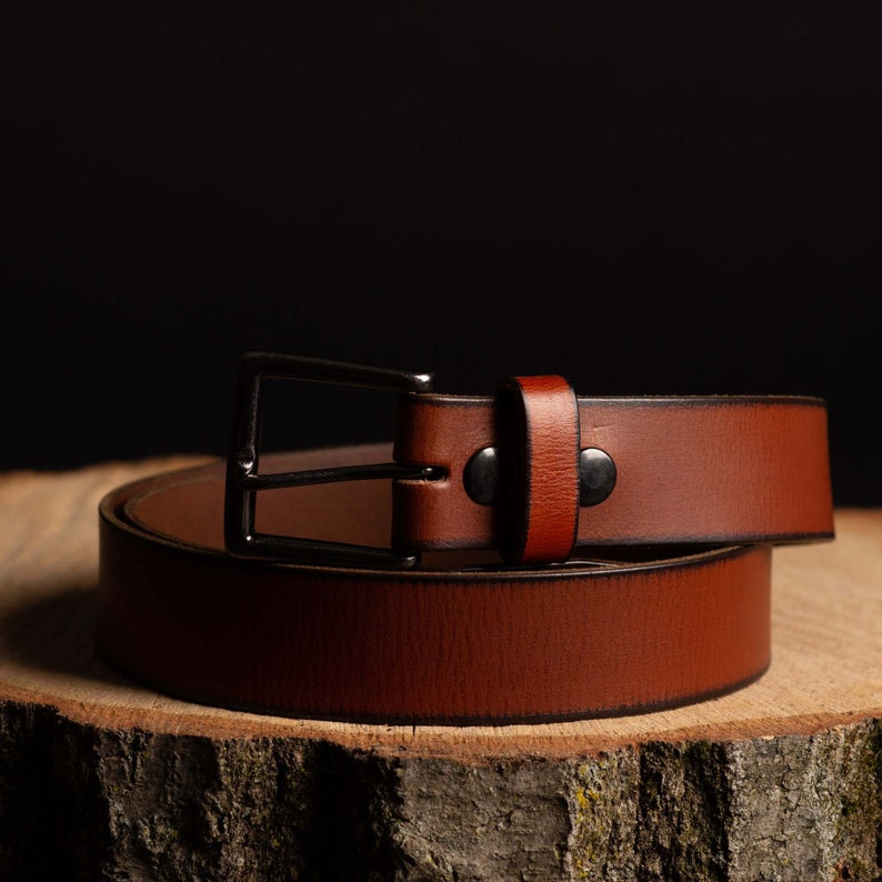 May include: A brown leather belt with a silver buckle. The belt is rolled up and sitting on a wooden surface.