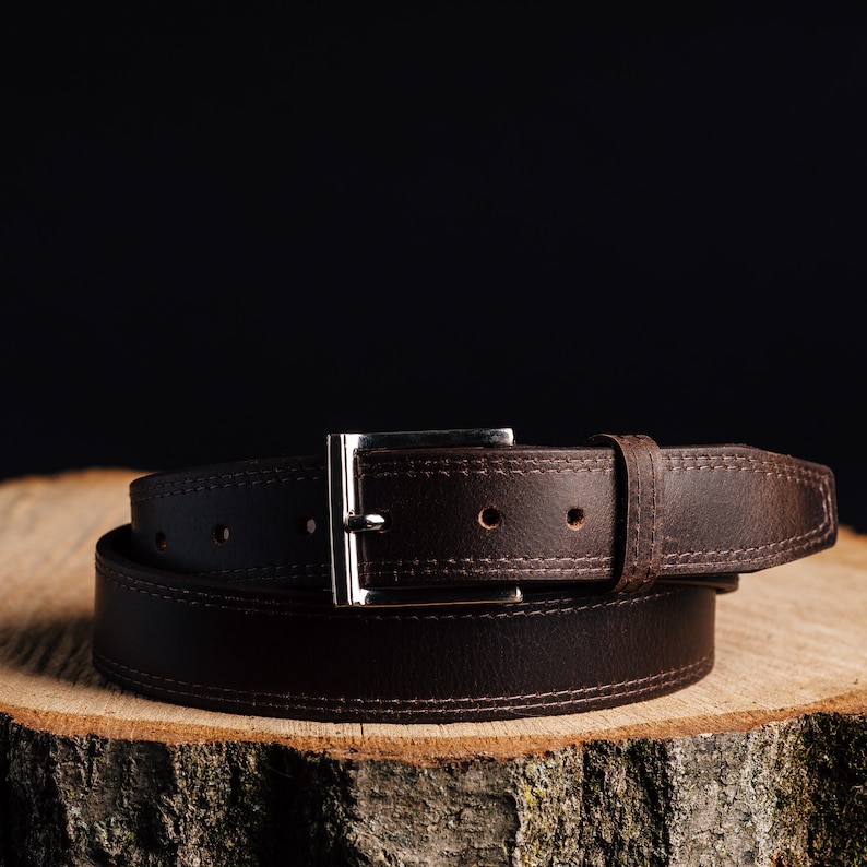May include: A brown leather belt with a silver buckle. The belt is on a wooden surface.