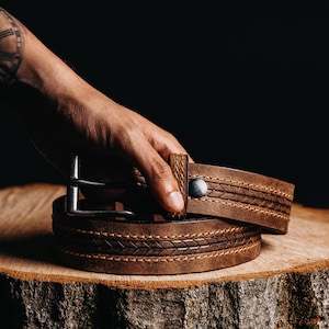 May include: A brown leather belt with a silver buckle and a decorative stitched pattern. The belt is displayed on a wooden surface against a dark background. The belt is approximately 1.5 inches wide.