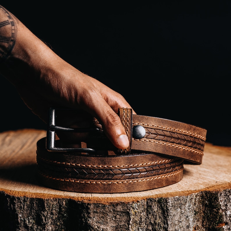 May include: A brown leather belt with a silver buckle and a decorative stitched pattern. The belt is displayed on a wooden surface against a dark background. The belt is approximately 1.5 inches wide.